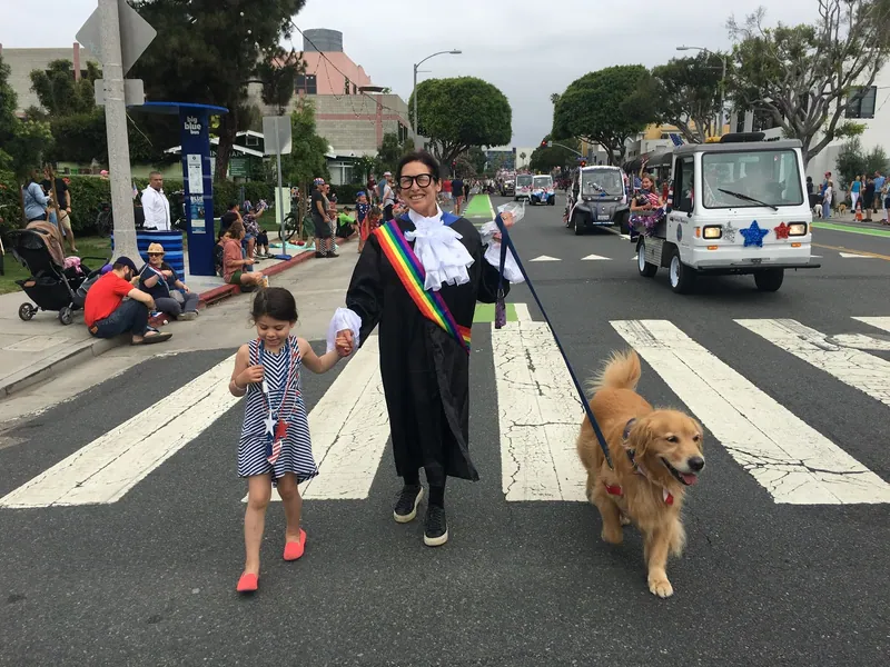 Sue Himmelrich in a judicial robe and rainbow sash walking in a Santa Monica Fourth of July parade with a young girl and a golden retriever.